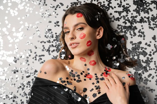 Top view of stylish woman with red lip prints lying near festive confetti and looking at camera on grey background - foto de stock