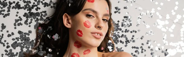 Top view of pretty brunette woman with red kiss prints looking at camera near shiny silver confetti on grey background, banner — Stock Photo