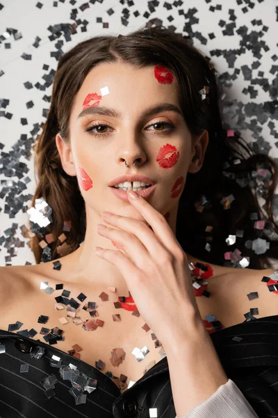 Top view of sensual woman with makeup and red kiss prints touching lip near silver confetti on grey background - foto de stock