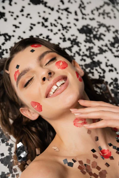 High angle view of cheerful woman with red lip prints and piercing lying with closed eyes near sparkling confetti on grey background - foto de stock