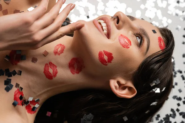 High angle view of smiling woman with red lipstick marks touching chin and looking away near sparkling confetti on grey background — Stock Photo
