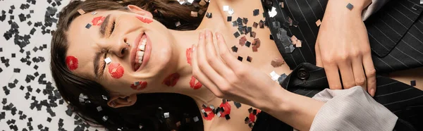 Top view of cheerful woman with red kiss prints lying with closed eyes near shiny confetti on grey background, banner — Stock Photo