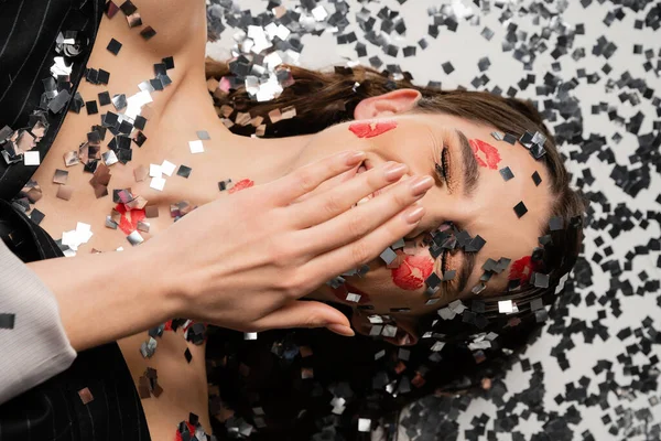 Top view of happy brunette woman with red kiss prints covering mouth with hand near silver confetti on grey background - foto de stock