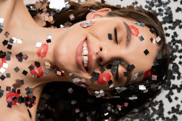 Top view of smiling woman with makeup and closed eyes lying near sparkling confetti on grey background — Stock Photo