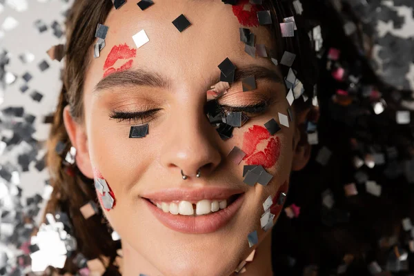 Close up portrait of young woman with red lip prints smiling with closed eyes near shiny confetti on grey background — Stock Photo