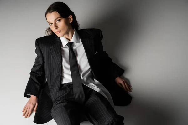 High angle view of stylish model in white shirt and black oversize suit looking at camera while sitting on grey background — Stock Photo