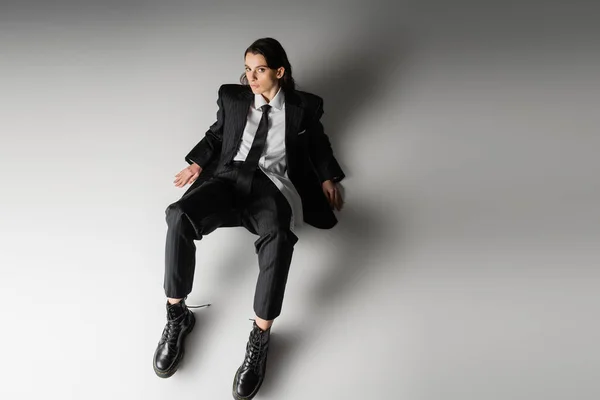 High angle view of brunette woman in black elegant suit and rough leather boots sitting and looking at camera on grey background — Stock Photo