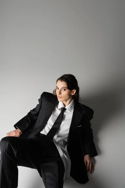 High angle view of brunette woman in black oversize suit and white shirt sitting and looking at camera on grey background — Foto stock