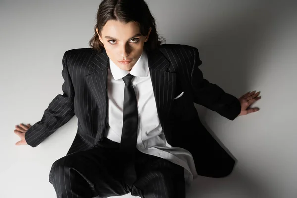 High angle view of young stylish woman in black suit and tie looking at camera while sitting on grey background — Stock Photo