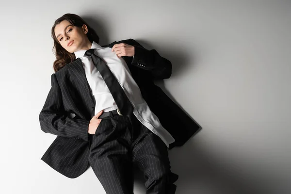 High angle view of brunette woman in white shirt and black striped suit lying with hand in pocket on grey background — Stock Photo