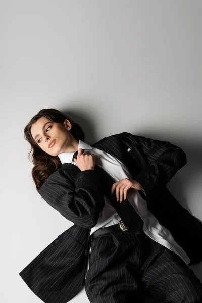 High angle view of brunette woman in black oversize suit touching tie and looking away while lying on grey background — Foto stock