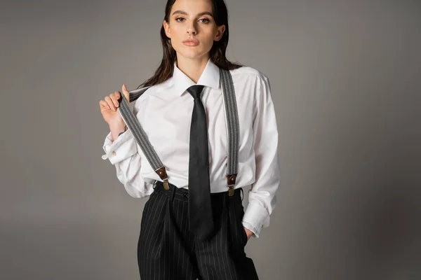 Pretty brunette woman in white shirt and tie standing with hand in pocket of black trousers with suspenders on grey background — Foto stock