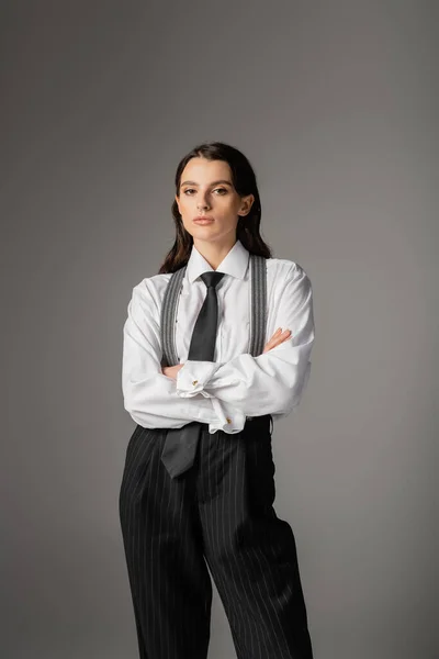 Stylish woman in white shirt and trousers with suspenders standing with crossed arms isolated on grey — Fotografia de Stock