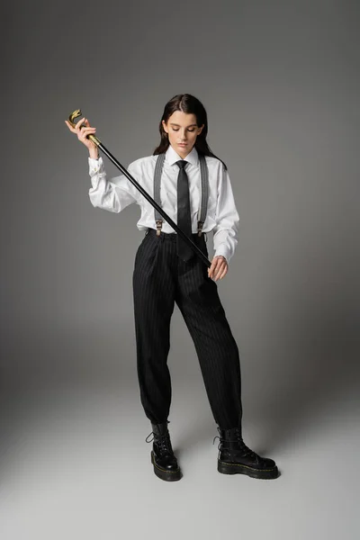 Full length of stylish woman in elegant oversize outfit and laced-up boots holding walking cane on grey — Stock Photo