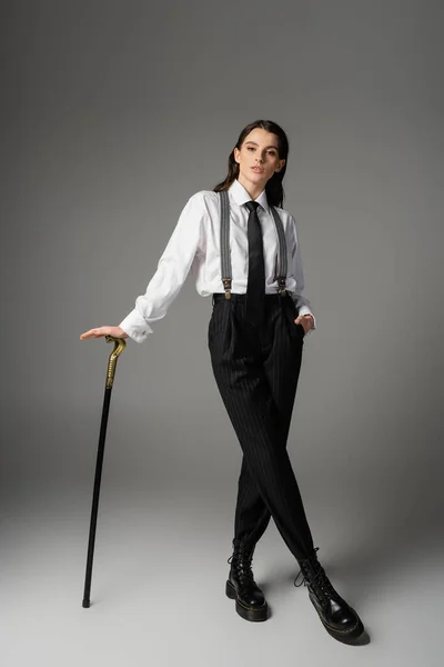 Full length of young woman in elegant clothes and rough leather boots posing with walking cane on grey background — Stockfoto