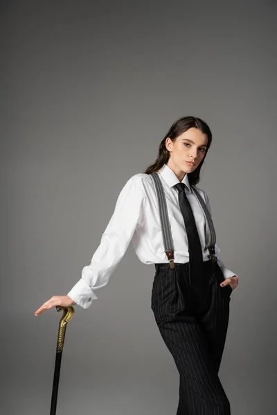 Pretty brunette model in white shirt and tie posing with walking cane and hand in pocket of black pants isolated on grey — Stock Photo