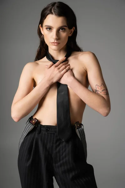 Shirtless tattooed woman in black tie and black striped pants covering breast and looking at camera isolated on grey - foto de stock
