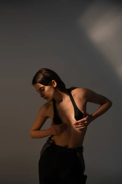 Brunette woman with shirtless body posing in black trousers and breast tape on grey background with lighting — Foto stock