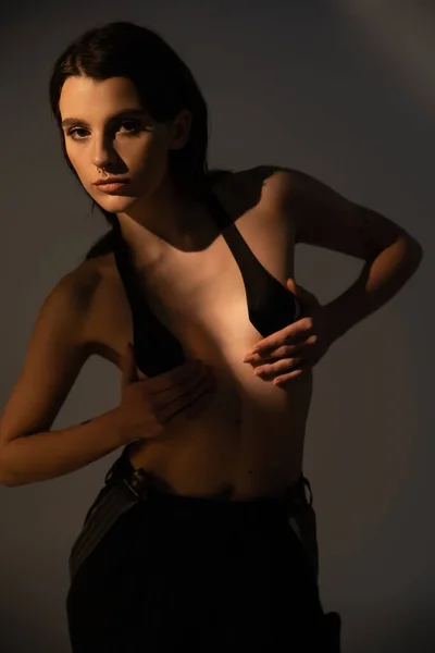 Молода без сорочки жінка в чорних штанях і грудній стрічці дивиться на камеру на сірому фоні з освітленням — стокове фото