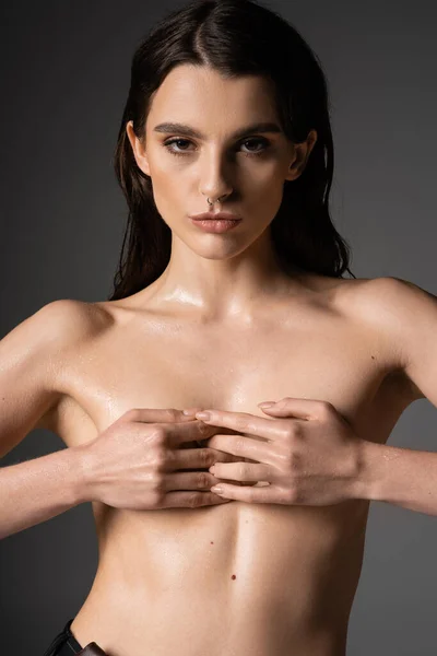 Seductive shirtless woman with wet body covering breast with hands and looking at camera on grey background — Stock Photo