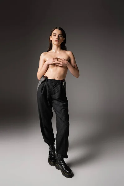 Shirtless woman with wet body covering breast with hands while posing in black pants and rough boots on grey — Foto stock