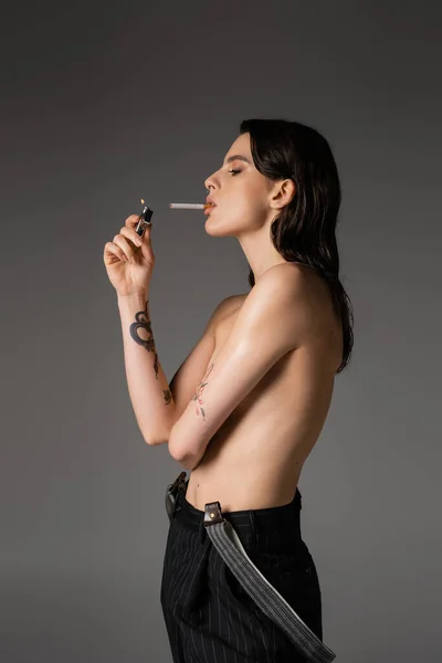 Side view of shirtless tattooed woman in black pants with suspenders lighting cigarette while standing isolated on grey - foto de stock