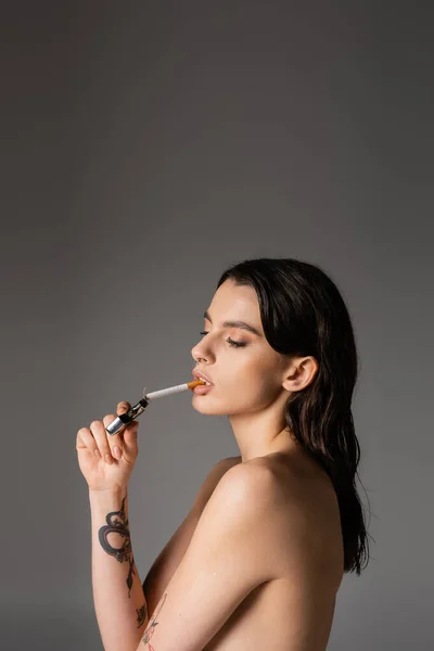Side view of shirtless tattooed woman lighting cigarette isolated on grey - foto de stock