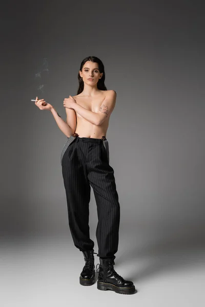 Full length of shirtless and sexy woman in black pants and rough boots posing with cigarette and looking at camera on grey — Foto stock