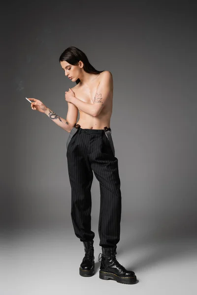 Full length of shirtless tattooed woman in trendy pants and laced-up boots covering breast and holding cigarette on grey - foto de stock