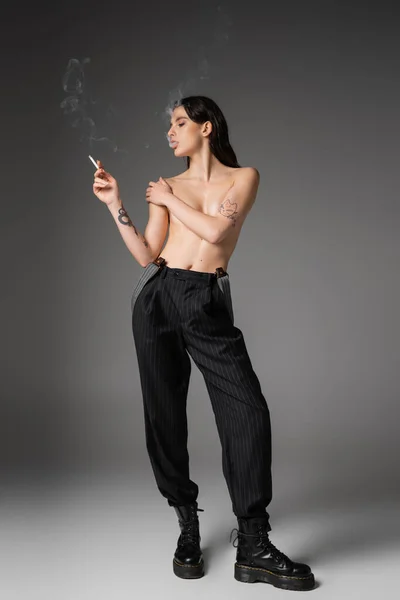 Volle Länge der hemdlosen verführerischen Frau in trendigen Hosen und groben Stiefeln, die Brust bedecken und Rauchen auf grauem Hintergrund — Stockfoto