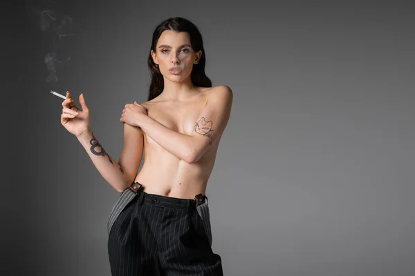 Sexy hemdlose Frau in schwarzer Hose, die die Brust bedeckt, während sie mit Zigarette auf grauem Hintergrund steht — Stockfoto