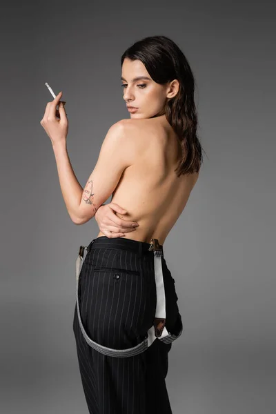 Без сорочки татуйована жінка в чорних штанях з підвісками позує з цигаркою ізольована на сірому — стокове фото