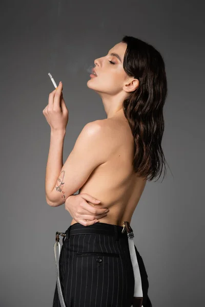 Вид збоку без сорочки і сексуальна жінка в чорних брюках з підтяжками, що стоять з цигаркою ізольовані на сірому — стокове фото