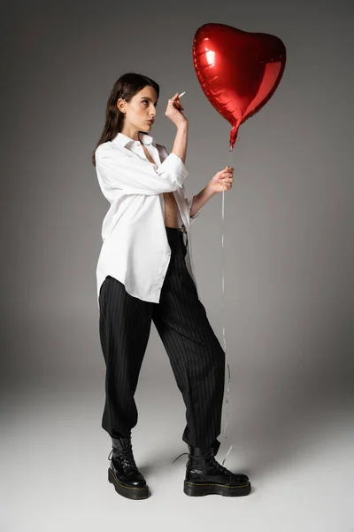 Full length of woman in white shirt and black pants with rough boots holding cigarette near red heart-shaped balloon on grey — Foto stock