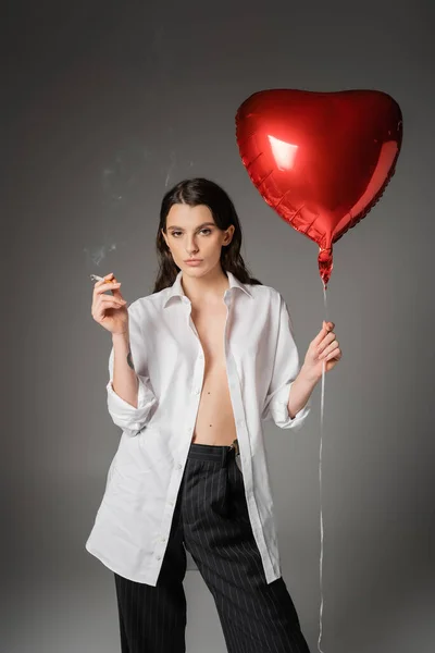 Trendy woman in white unbuttoned shirt holding cigarette and red heart-shaped balloon while looking at camera on grey — Stock Photo