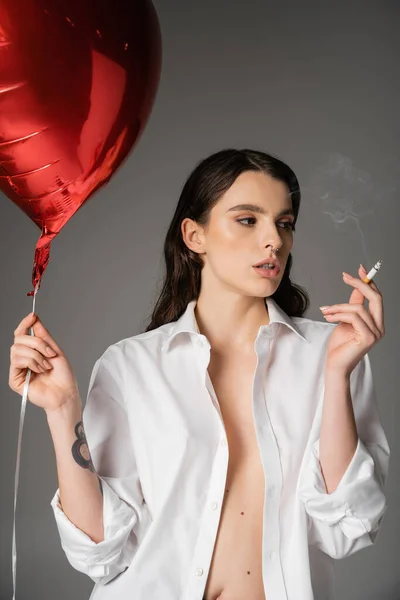Sexy woman in white unbuttoned shirt holding cigarette and red heart-shaped balloon while looking away isolated on grey — Fotografia de Stock