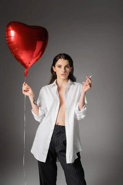 Stylish woman with red heart-shaped balloon and cigarette posing in white unbuttoned shirt on grey background — Fotografia de Stock