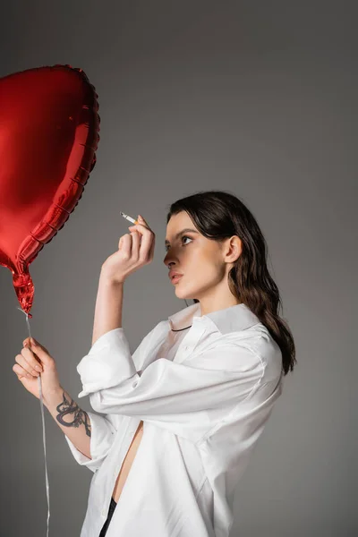 Mujer tatuada de moda en camisa blanca de pie con cigarrillo y globo rojo en forma de corazón aislado en gris - foto de stock