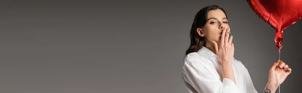 Brunette woman in white shirt smoking while holding red balloon isolated on grey, banner — Stock Photo