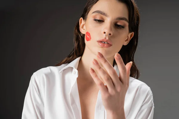 Молода брюнетка з червоним поцілунком друкує на обличчі, дивлячись на руку на сірому фоні — стокове фото