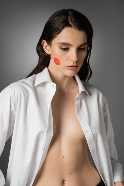 Сексуальная женщина с красным отпечатком губы на лице, позирующая в белой расстегнутой рубашке на сером — стоковое фото