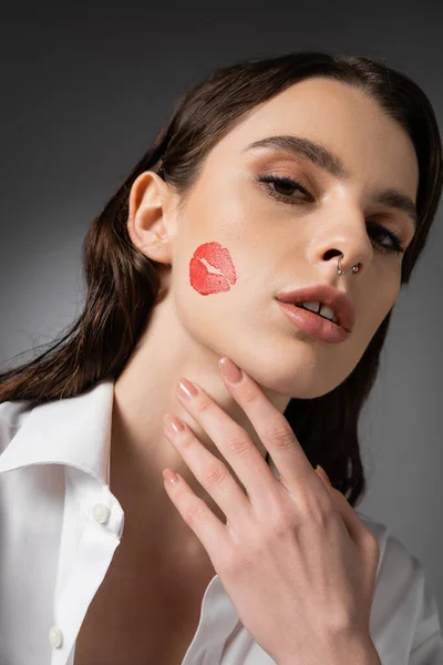 Portrait of brunette woman with red kiss print on face touching neck and looking at camera on grey background — Foto stock