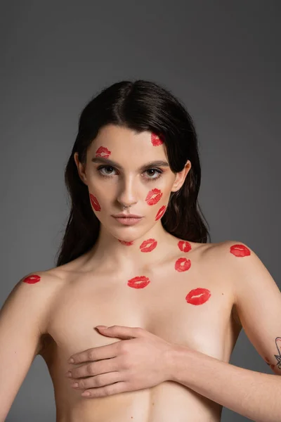 Sexy shirtless woman with red kiss prints on body and face covering breast with hand and looking at camera isolated on grey — Fotografia de Stock