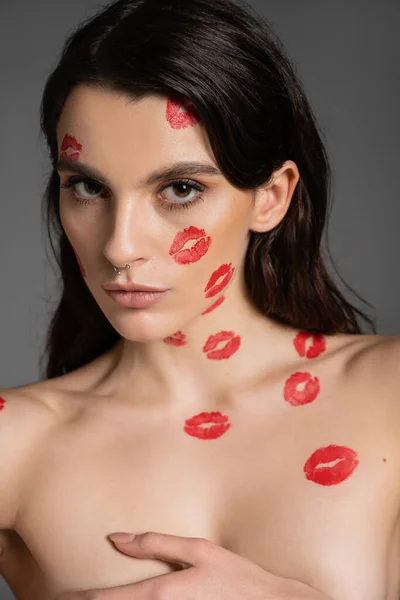 Сексуальна без сорочки жінка з червоними відбитками губ, що покривають груди рукою, дивлячись на камеру ізольовано на сірому — стокове фото