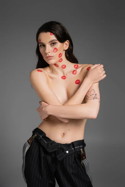 Sexy tattooed woman with red lip prints on shirtless body covering bust with crossed arms and looking at camera isolated on grey — Stockfoto