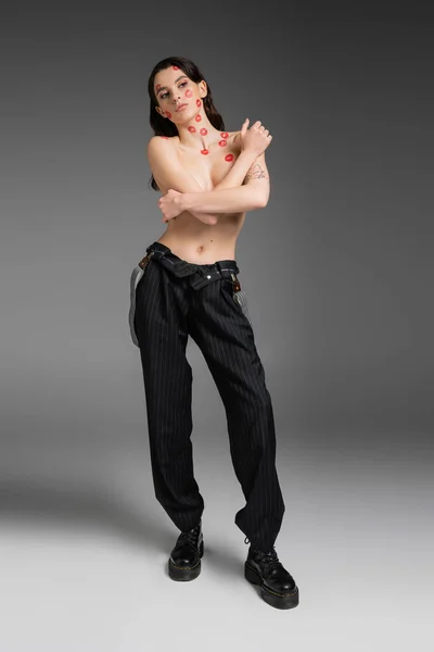 Full length of sexy shirtless woman with red lipstick marks on face and body posing in black pants and rough boots on grey — Stock Photo