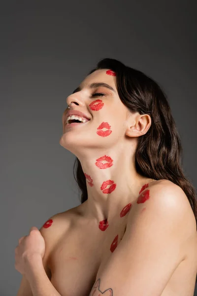Young brunette woman with red kiss prints on face and shirtless body laughing with closed eyes isolated on grey — Foto stock