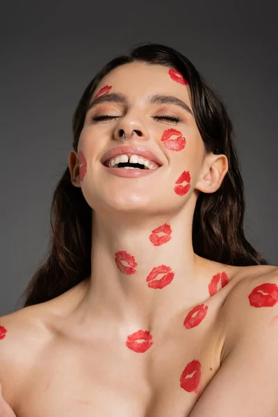 Excited woman with bare shoulders and red lipstick marks on body and face laughing with closed eyes isolated on grey — Foto stock