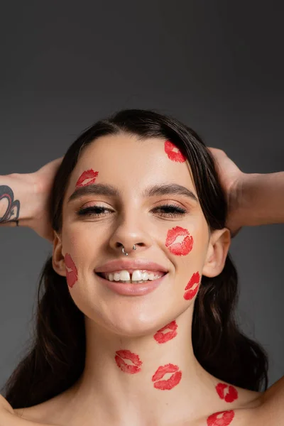 Портрет веселой татуированной женщины с красными поцелуями на лице, касающимися волос и улыбающейся в камеру, изолированную на сером — стоковое фото
