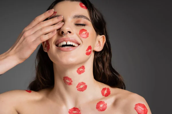 Cheerful woman with red lip prints on face and bare shoulders covering eye with hand isolated on grey — Foto stock